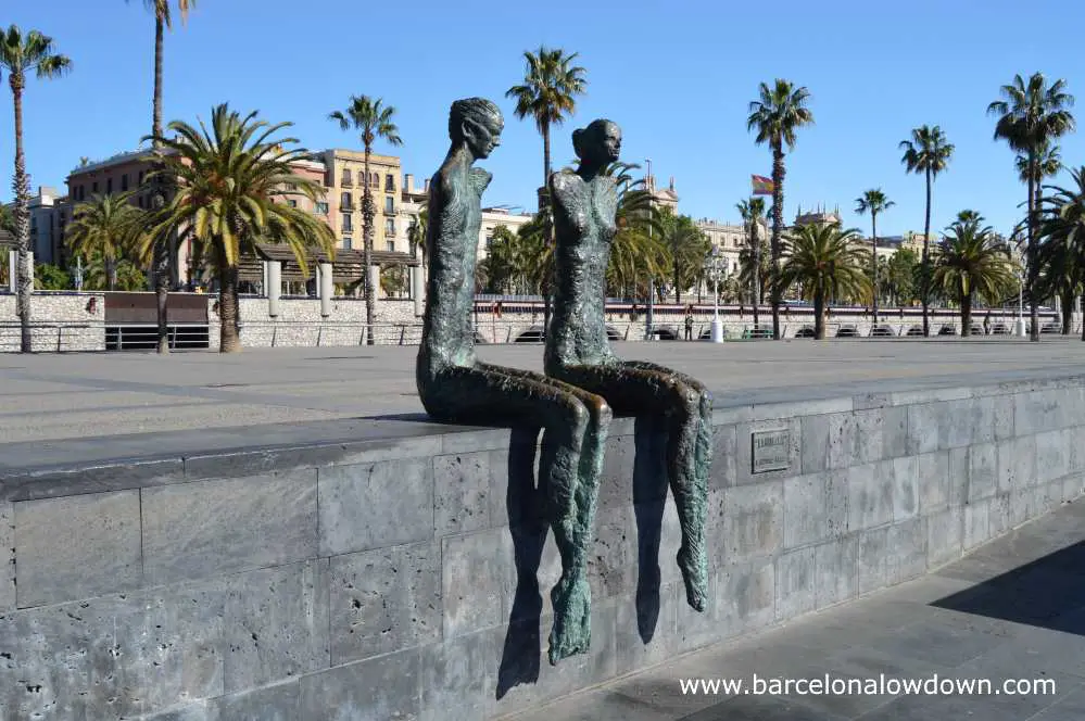 The couple statue by Lauturo Díaz sit alone on a wall in front of old buildings beside Barcelona's historic old port the Port Vell