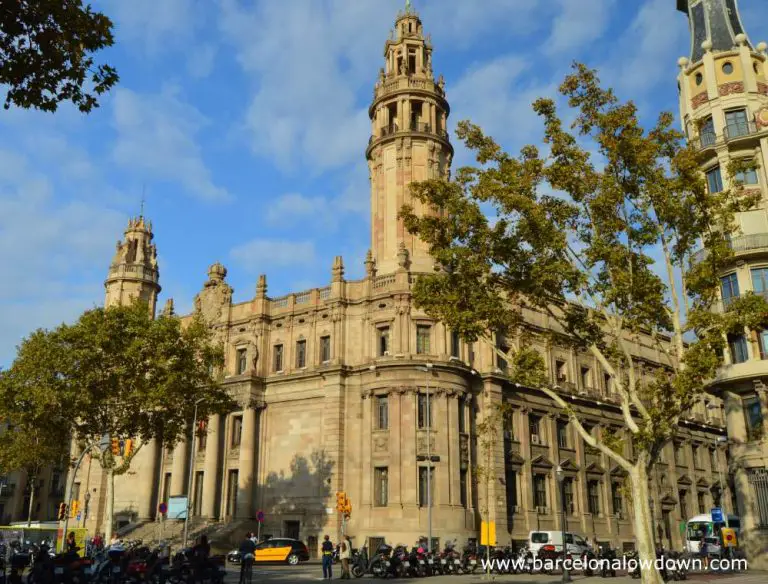 The exterior of Barcelona central post office seen from the other side of Via Laitana. There are two trees in the foreground and a line of parked motorbikes on the kerb.