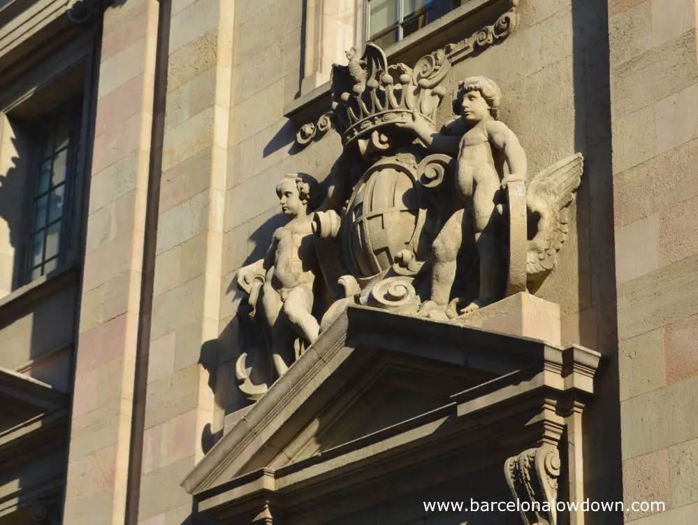 Cherubs holding a crown with a bat on it above the Barcelona coat of arms. Carved details above a door at the side of Barcelona central post office
