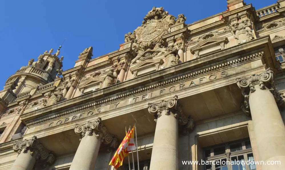 Close-up of the carvings above the main entrance to Barcelona Central Post Office. There is a large coat of arms and several stone statues above the greek style entrance way with decorated columns. At the bottom of the photo there are two flags one is the Catalan flag the other is the flag of Barcelona.