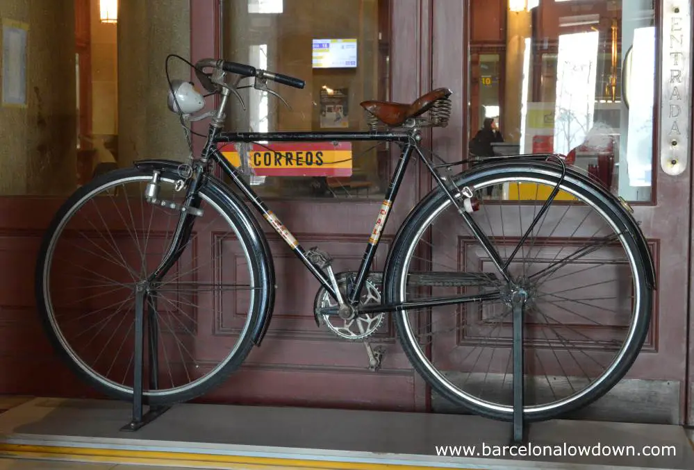 One of the two black vintage postman's bikes located just inside the entrance of Barcelona Central Post Office. The bike is standing in front of brown doors with windows through which you can see the interior of the post office.