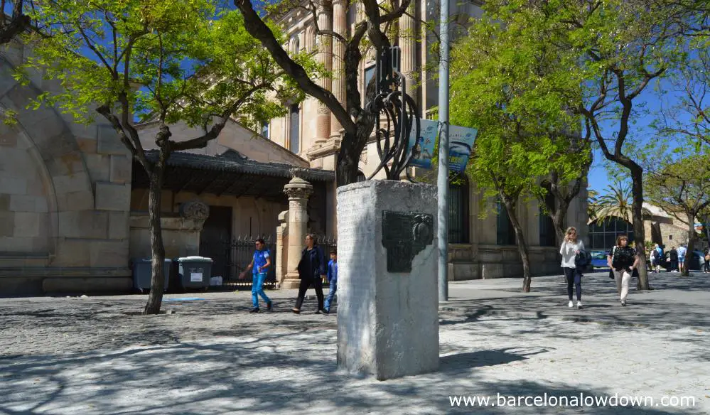 3.5m stone and iron monument in front of the old customs house in Barcelona. The monument is surrounded by trees and a few people on a sunny spring day.