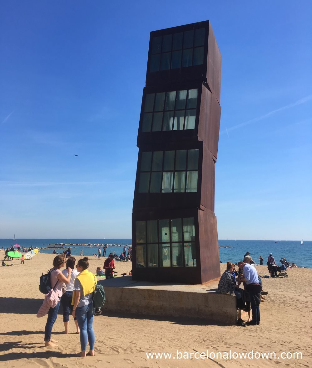 Photo of Barcelona's main beach La Barcelonaeta taken on a sunny Saturday morning in February. In the centre of the photo is the rusty steel wounded star monument by Rebecca Horm surrounded by people who are enjoying the winter sunshine barefoot in the sand. A couple of surfers carry their SUP boards to the sea. Several yachts can be seen sailing in the background.