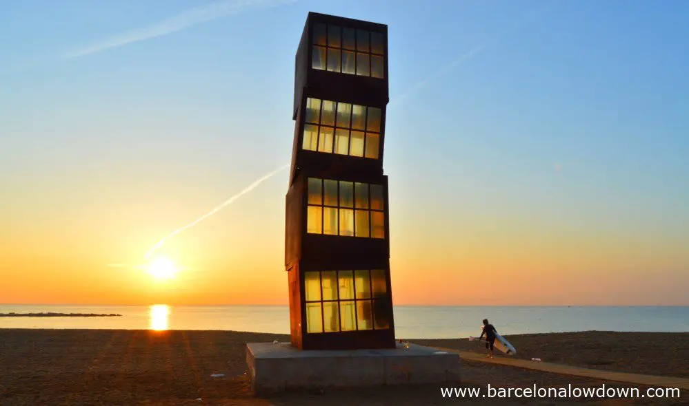 The wounded star statue at sunrise by Rebecca Horn. Photo of Barcelona beach, the early morning sunshine is shining through windows of thye rusty iron block statue staining them yellow. A lone paddlesurfer is walking across the sand carring their board towards the sea for an early morning surf session,