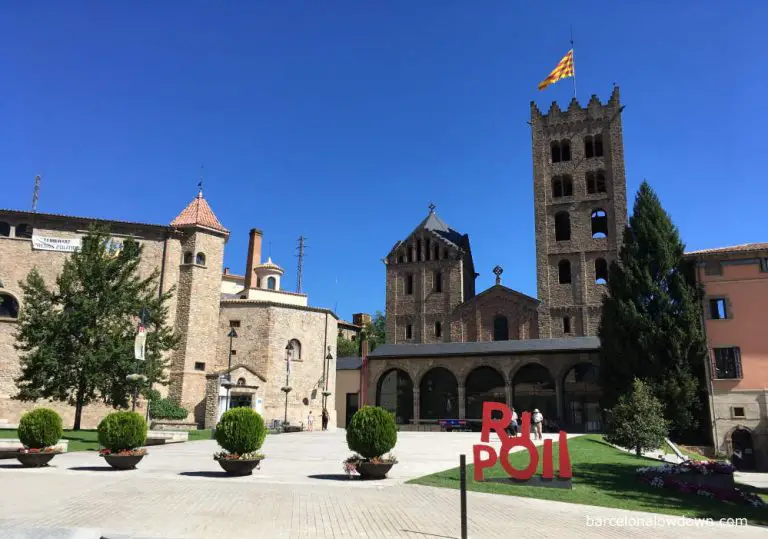 The plaza in front of the Monastery of Santa Maria de Ripoll on a sunny day. A yellow and red senyera flag flys at the top of the bell tower of the monastery.