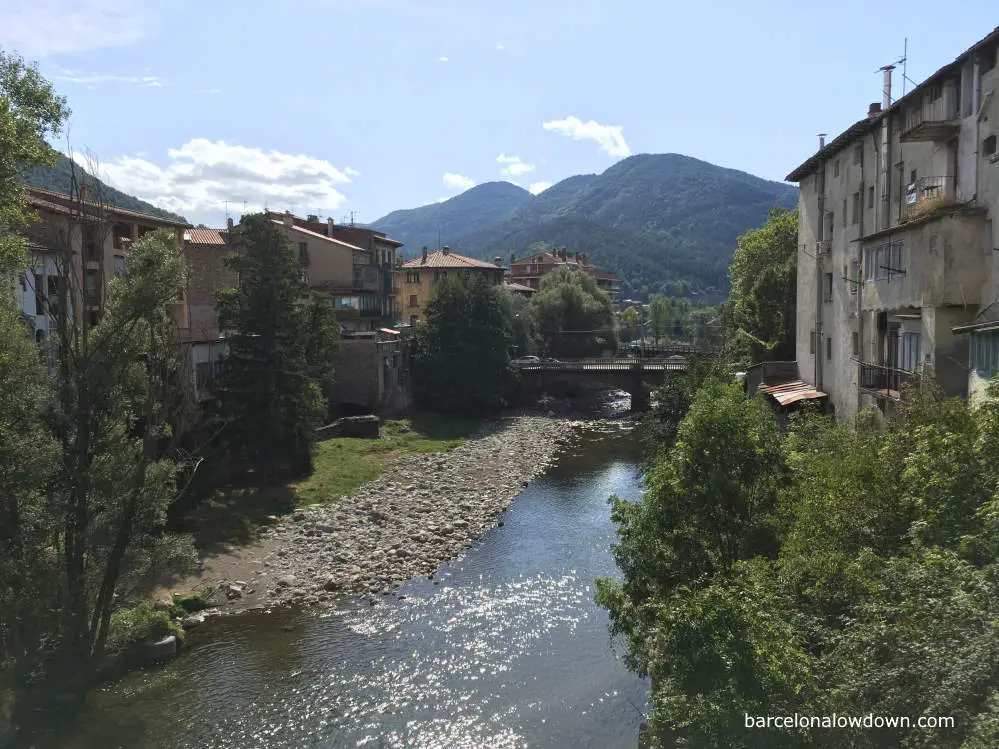 The River Freser as it passes through Ripoll with trees and old buildings either side and green mountains in the background.
