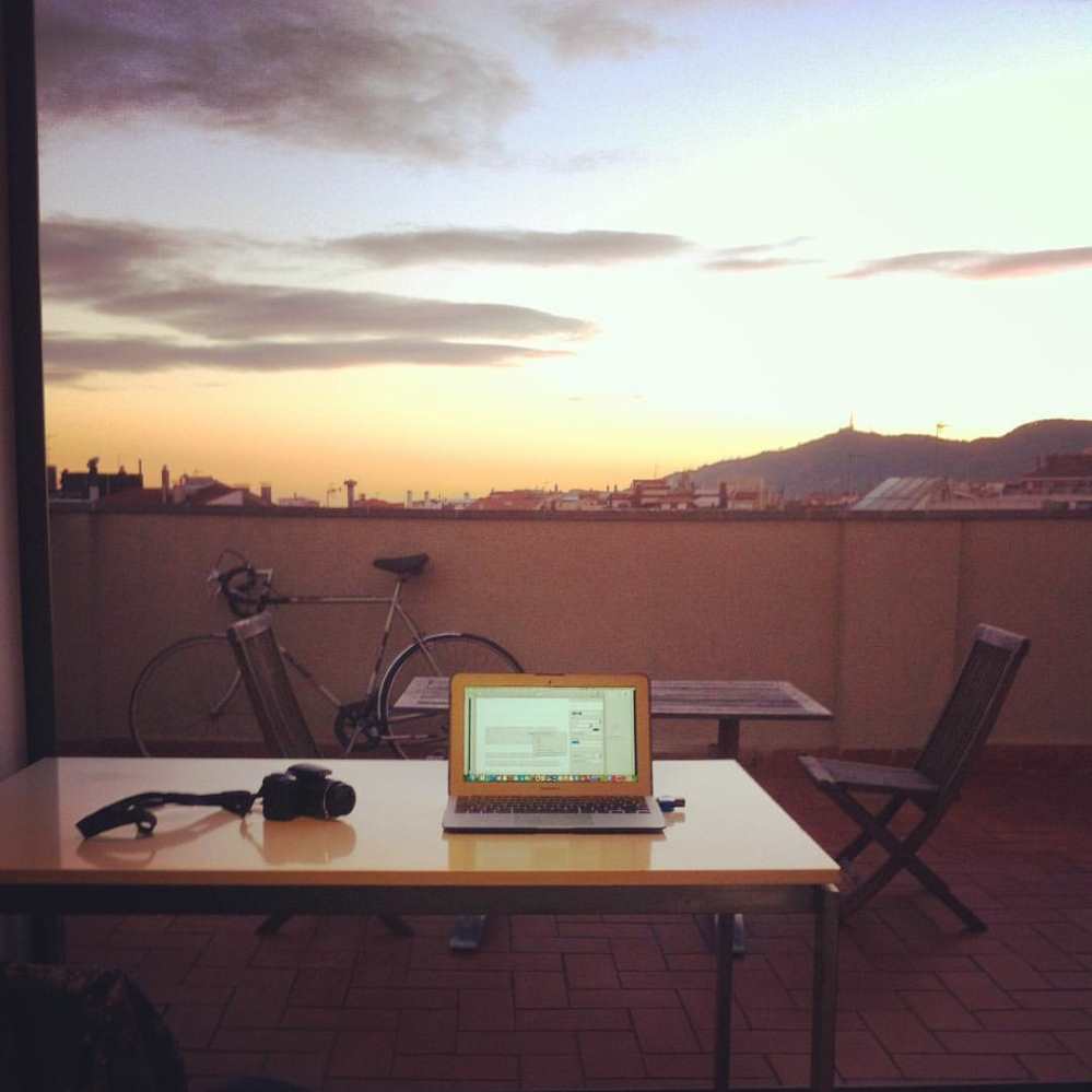 Ben Holbrook's desk on a roof terrace in Barcelona, his laptop and camera are on the desk and the sun is setting in the background