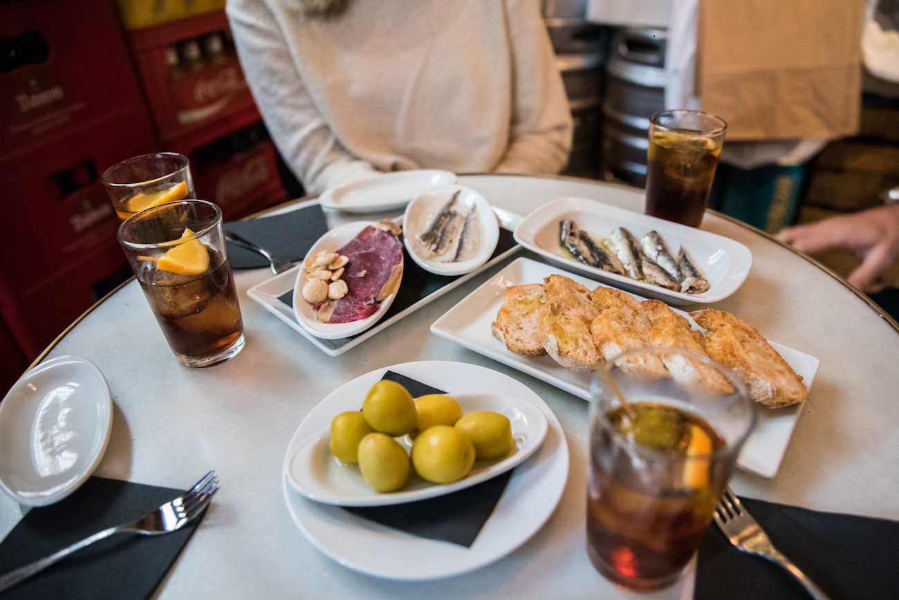 A table in a tapas bar, there are four glasses of vermouth, some olives, some bread, Spanish ham and sardines on plates